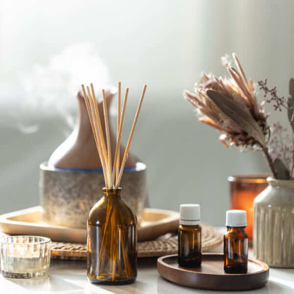 An aromatherapy setup featuring a brown glass bottle with wooden reed sticks, two small amber essential oil vials on a wooden tray, and a ceramic ultrasonic diffuser releasing mist in the background.