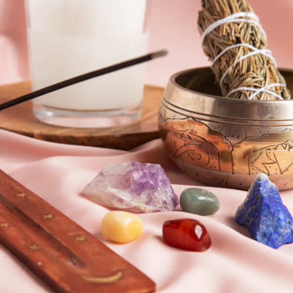 Une collection d'objets de bien-être spirituel comprenant plusieurs cristaux bruts et polis, un porte-encens en bois et un bol chantant en laiton contenant un bouquet de sauge séchée sur une surface en tissu rose.