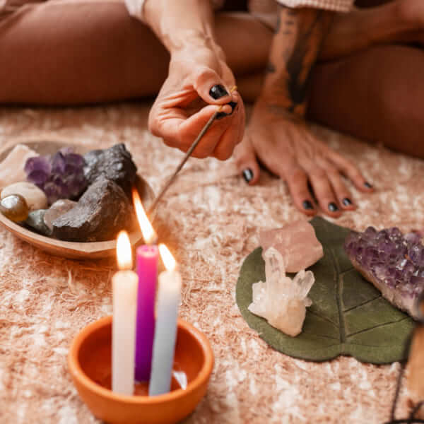 Un gros plan d'une main tatouée allumant un bâton d'encens au-dessus de trois petites bougies allumées, entourée de divers cristaux bruts comme de l'améthyste et du quartz sur un tapis texturé.